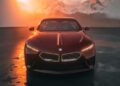 A red BMW Shooting Brake is parked on a wet surface, framed by snow-capped mountains and an orange sunset in the background.
