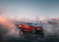 A red BMW Speedtop V8 sports car is parked on a wet surface with mountains and clouds in the background at sunrise or sunset.