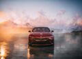 A red BMW Shooting Brake is parked on a reflective surface, with snowy mountains and a colorful sky in the background.
