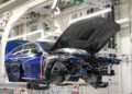 Partially assembled blue BMW M4 CS with open hood on an automotive production line, surrounded by machinery and overhead equipment, highlighting the precision of luxury manufacturing.
