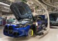 A blue BMW M4 CS with its hood open is being assembled on a production line in an automotive factory.