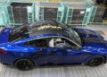 A blue BMW M4 CS sports car with black wheels is parked inside a factory setting, surrounded by green metal storage racks and safety barriers, showcasing a touch of bespoke luxury.
