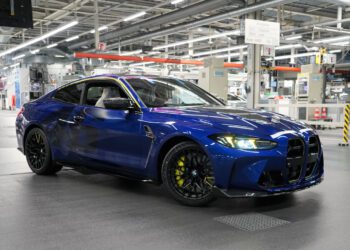 A blue BMW M4 CS coupe parked inside an automotive manufacturing facility, surrounded by industrial equipment and bright overhead lighting, exudes bespoke luxury and engineering excellence.