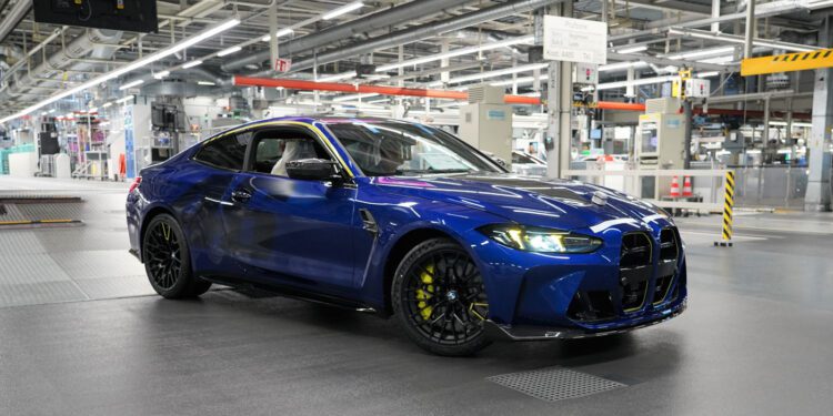 A blue BMW M4 CS coupe parked inside an automotive manufacturing facility, surrounded by industrial equipment and bright overhead lighting, exudes bespoke luxury and engineering excellence.