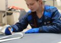 A woman in a blue uniform and gloves works at a table, meticulously writing or drawing on material for a bespoke luxury project in an indoor workspace.