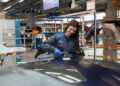 A woman in a blue uniform uses a polishing tool on the hood of a bespoke luxury BMW M4 CS inside an automotive manufacturing facility.