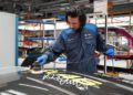 A worker in a BMW Group uniform uses a polishing machine on the hood of a BMW M4 CS with a large white signature and yellow markings, highlighting the bespoke luxury of the exclusive VR46 Edition in an automotive workshop.