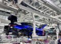 Partially assembled cars, including the bespoke luxury BMW M4 CS VR46 edition, move along an automated production line in a modern automotive factory, with machinery overhead and workers in the background.