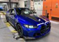 A blue BMW M4 CS coupe on a factory testing platform inside an automotive facility, with machinery and safety signs visible in the background, highlights bespoke luxury engineering.