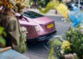 A pink Rolls-Royce Spectre with the license plate HT14 YOM is parked on a street in Chelsea, London, partially obscured by a person holding flowers and various colorful floral arrangements.