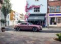 A pink Rolls-Royce Spectre coupe glides along a quiet Chelsea London street, lined with colorful buildings, one adorned with pink flowers.