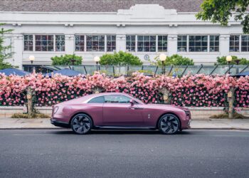 A mauve-colored Rolls-Royce Spectre luxury coupe is parked on a street in Chelsea, London, in front of a building with large windows and a pink flower-covered fence.