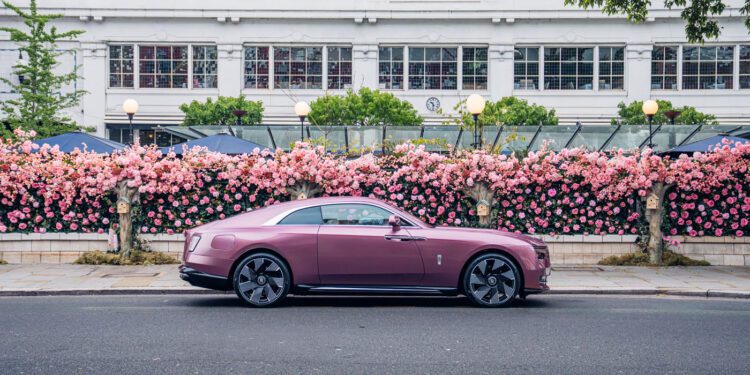 A mauve-colored Rolls-Royce Spectre luxury coupe is parked on a street in Chelsea, London, in front of a building with large windows and a pink flower-covered fence.