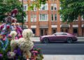 A pink Rolls-Royce coupe, possibly the Spectre, is parked on a Chelsea London street across from a decorative flower display shaped like a poodle and a person in a hat, with brick apartment buildings in the background.
