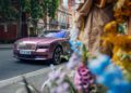 A pink Rolls-Royce Spectre is parked on a Chelsea London street, partially obscured by colorful flowers in the foreground.