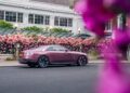 A mauve Rolls-Royce Spectre coupe is parked on the street in Chelsea London beside a row of pink flowering bushes, with a white building in the background and blurred pink flowers in the foreground.
