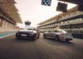 Two sports cars, including the 2026 M2 CS boasting 523HP, are parked side by side on a racetrack near the pit lane, surrounded by grandstands and stadium structures.