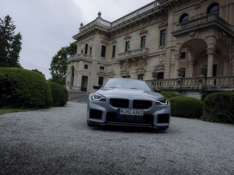 A silver BMW M2 CS sports car is parked on a gravel driveway in front of an ornate, historic mansion with stone columns and manicured bushes, looking ready for its world premiere.
