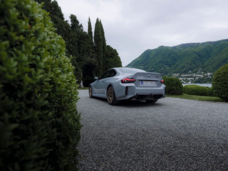 A light blue BMW M2 CS coupe is parked on a gravel driveway surrounded by green shrubs, with hills and a cloudy sky in the background.