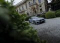 A silver BMW M2 CS sports car parked on a gravel driveway in front of an ornate, historic building with manicured bushes.