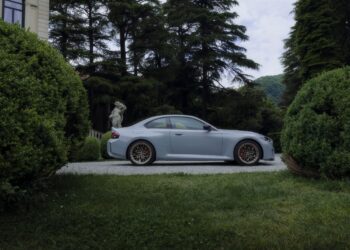 A silver BMW M sports coupe parked on a gravel driveway, surrounded by manicured bushes and tall trees, with a stone statue in the background.
