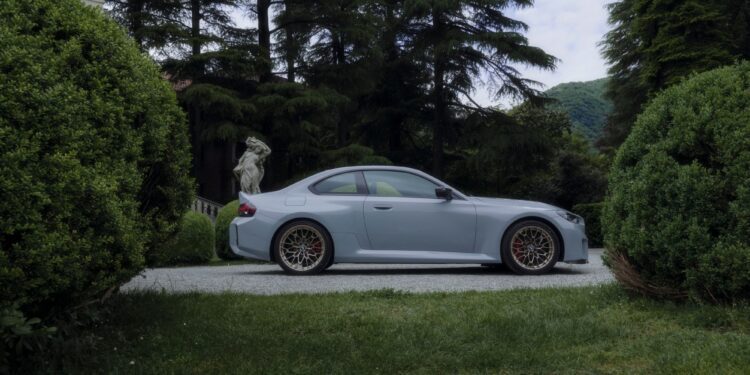 A silver BMW M sports coupe parked on a gravel driveway, surrounded by manicured bushes and tall trees, with a stone statue in the background.