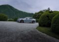A silver BMW M2 CS sports car is parked on a gravel driveway, framed by green manicured bushes and tree-covered hills in the background under a cloudy sky.