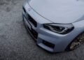 Close-up of the front end of a light gray BMW M2 CS, showing the grille, headlights, and part of the hood, parked on a gravel surface—a striking look for this BMW M series world premiere.