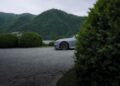 A silver BMW M2 CS is partially visible behind trimmed bushes, with green hills and a lake in the background under a cloudy sky—a perfect scene for a world premiere.