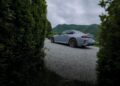 A silver BMW M2 CS sports coupe with bronze wheels is parked on a gravel driveway, framed by green hedges, with green hills and a cloudy sky in the background—a scene worthy of a world premiere.