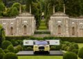 A yellow Rolls-Royce Phantom is displayed on a black platform in front of an ornate garden palace, reminiscent of Villa d'Este, with manicured hedges and decorative stonework evoking a scene from Goldfinger.