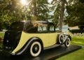 A vintage yellow and black Rolls-Royce Phantom with whitewall tires is displayed outdoors on a platform at Villa d'Este, surrounded by trees and greenery, evoking the elegance of Goldfinger-era classics.
