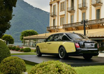 A two-tone Rolls-Royce Phantom is displayed on a platform in a landscaped garden near a large building at Villa d’Este, with mountains and a lake in the background, evoking the style of Goldfinger.