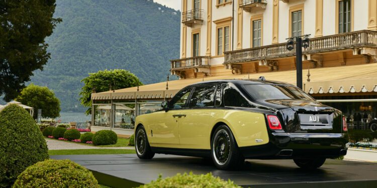 A two-tone Rolls-Royce Phantom is displayed on a platform in a landscaped garden near a large building at Villa d’Este, with mountains and a lake in the background, evoking the style of Goldfinger.