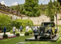 A classic black and yellow Rolls-Royce Phantom, reminiscent of Goldfinger, is displayed on a platform in a manicured garden at Villa d'Este, with people strolling and historic buildings in the background.