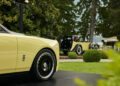Two classic yellow and black cars, including a Rolls-Royce Phantom reminiscent of Goldfinger, are parked outdoors on a landscaped lawn at Villa d'Este, with trees, bushes, and garden seating visible in the background.