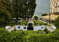 A manicured garden at Villa d'Este features cars, including a Rolls-Royce Phantom, parked on a gravel path, photo displays on stands, and a large building overlooking the lake and mountains—evoking the elegance of a Goldfinger scene.