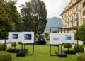 A black Rolls-Royce Phantom is parked on a lawn between two display stands showing car photographs at Villa d'Este, with a large building and mountains in the background, evoking the elegance of Goldfinger.