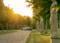 A silver Pagani hypercar is parked on a cobblestone path lined with trees and classical statues at sunset, capturing the spirit of Motor Valley Fest.