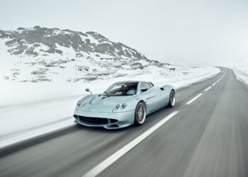A silver Pagani hypercar drives on an empty, snow-lined mountain road under an overcast sky.