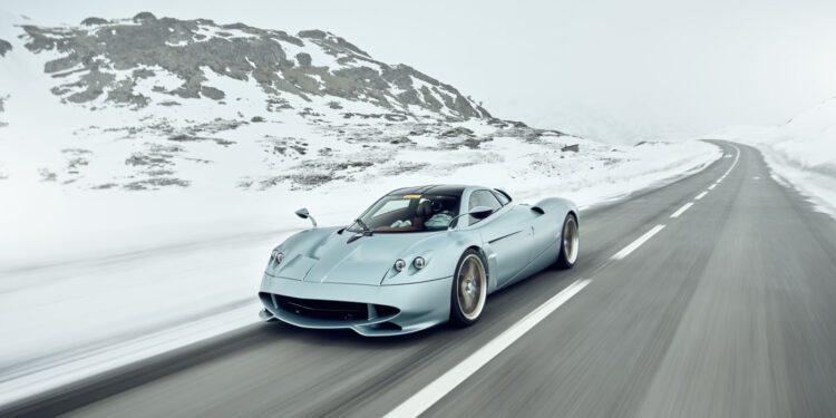 A silver Pagani hypercar drives on an empty, snow-lined mountain road under an overcast sky.