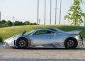 A silver Pagani Huayra Epitome supercar is parked on a cobblestone surface near Villa d'Este, with grass, trees, and streetlights in the background on a clear day.