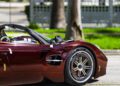 A close-up side view of a luxury sports car, possibly a Pagani, parked on a street with the front wheel and fender in focus—green trees in the background evoke the atmosphere of Motor Valley Fest celebrating hypercars.