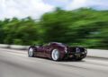 A maroon Pagani hypercar driving at speed on a highway, with blurred green trees and sky in the background.