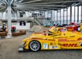 Several brightly colored Porsche 963 race cars, inspired by Roger Penske’s legendary race team, are displayed inside a modern showroom with large windows, metal beams, and a seating area.