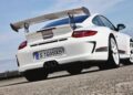 A white sports car— the last production Porsche with a Mezger flat-six engine, the GT3 RS 4.0— is parked on a paved surface, viewed from the back left side with its large rear spoiler in full display.