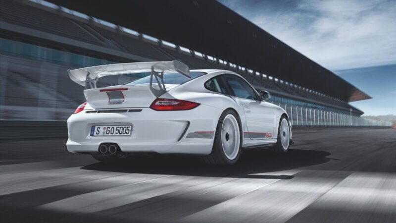 A white sports car— the last production Porsche with a naturally-aspirated Mezger flat-six engine—speeds down a racetrack, with empty grandstands visible in the background.