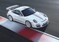 A white Porsche sports car with red accents—known as the last production Porsche with a naturally-aspirated Mezger flat-six engine—is driving on a racetrack, positioned near the red and white curbing.