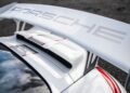 Close-up of a white Porsche sports car’s rear wing with the Porsche logo, showcasing the aerodynamic design of the last production Porsche with a naturally-aspirated Mezger flat-six engine against dark asphalt.