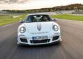 A white Porsche sports car, the last production Porsche with a naturally-aspirated Mezger flat-six engine, speeds down a racetrack, captured from the front with motion blur on the road and background.